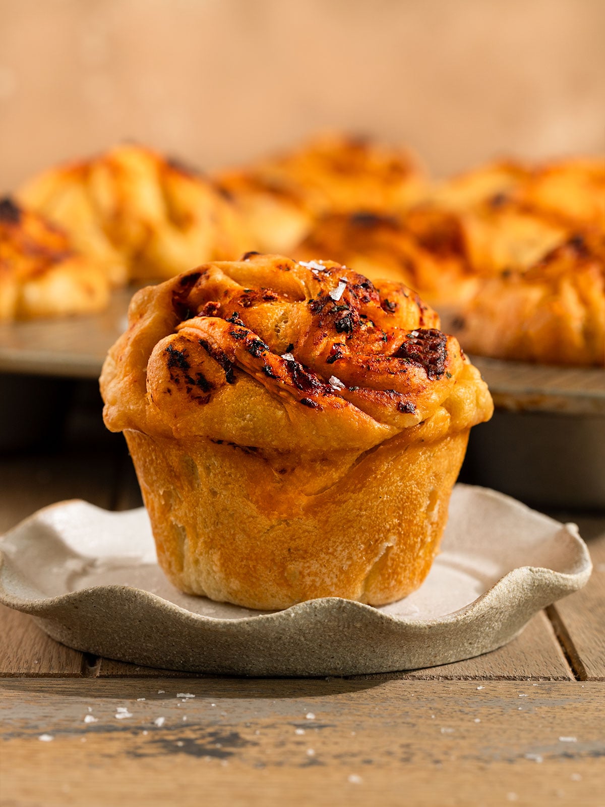 focaccia muffin on plate topped with a tomato garlic oil and flaky sea salt in front of tray of more muffins.
