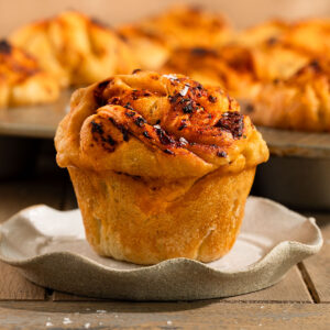 focaccia muffin on plate topped with a tomato garlic oil and flaky sea salt in front of tray of more muffins.