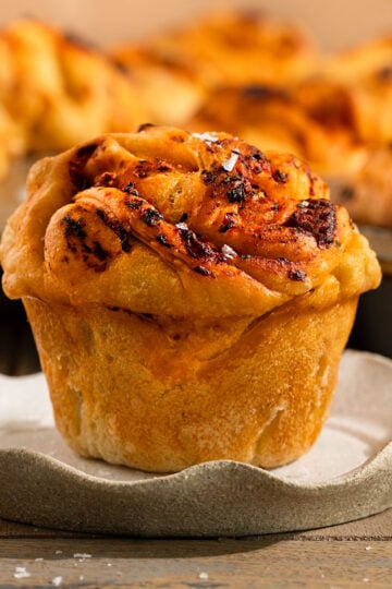 focaccia muffin on plate topped with a tomato garlic oil and flaky sea salt in front of tray of more muffins.