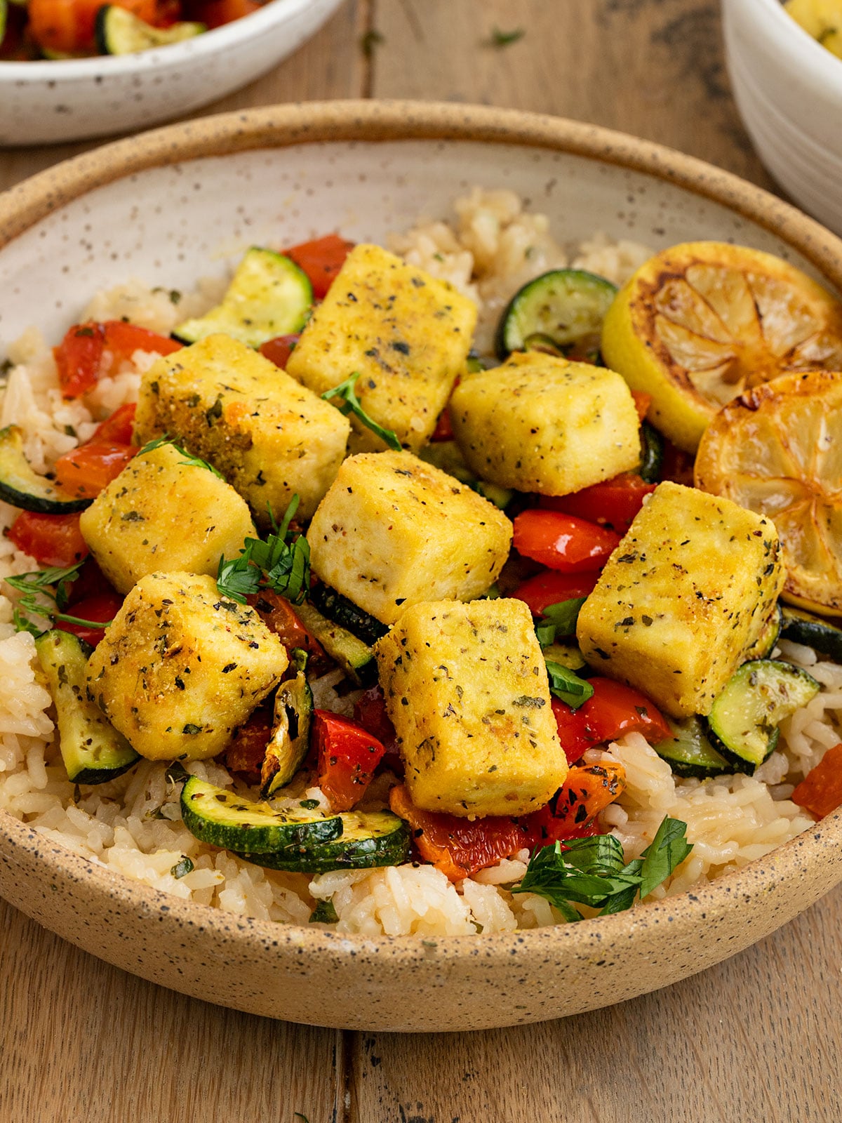 bowl filled with rice, red peppers, zucchini, pan fried tofu squares, and charred lemon slices topped with fresh parsley.