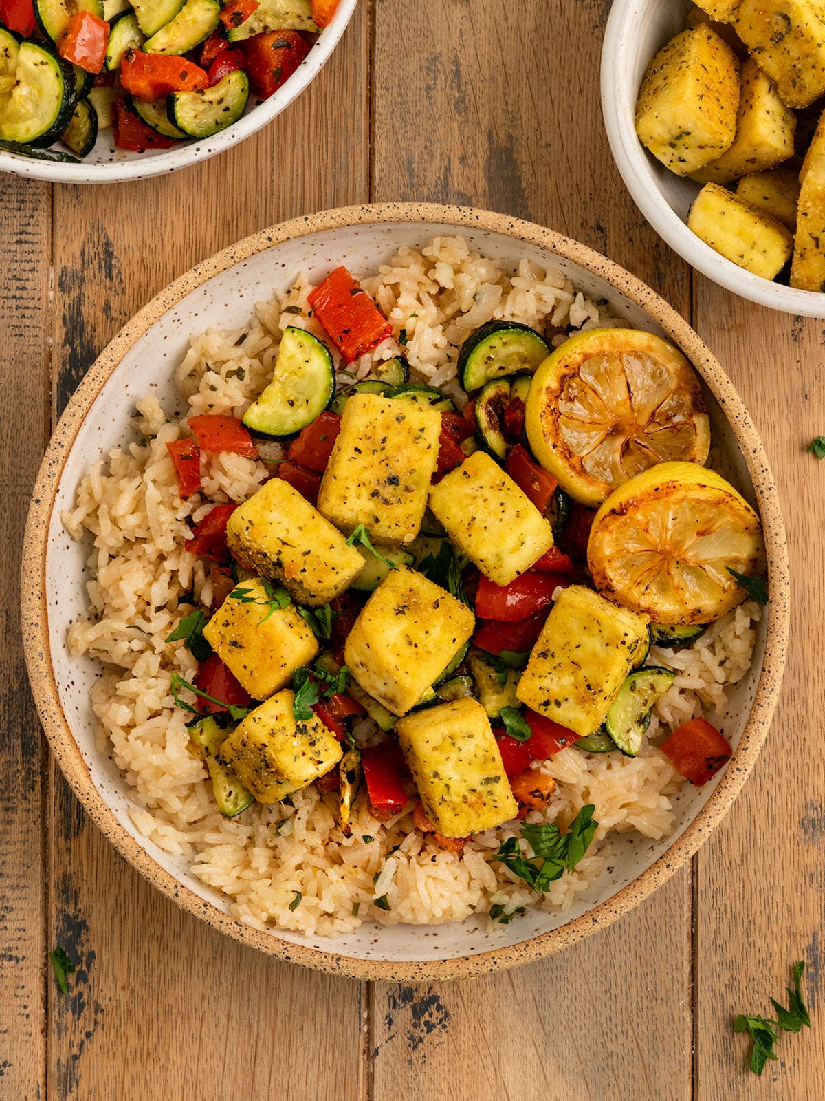bowl filled with rice, red peppers, zucchini, pan fried tofu squares, and charred lemon slices, a spoon, and topped with fresh parsley.
