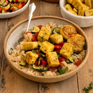 bowl filled with rice, red peppers, zucchini, pan fried tofu squares, and charred lemon slices, a spoon, and topped with fresh parsley.