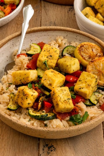 bowl filled with rice, red peppers, zucchini, pan fried tofu squares, and charred lemon slices, a spoon, and topped with fresh parsley.