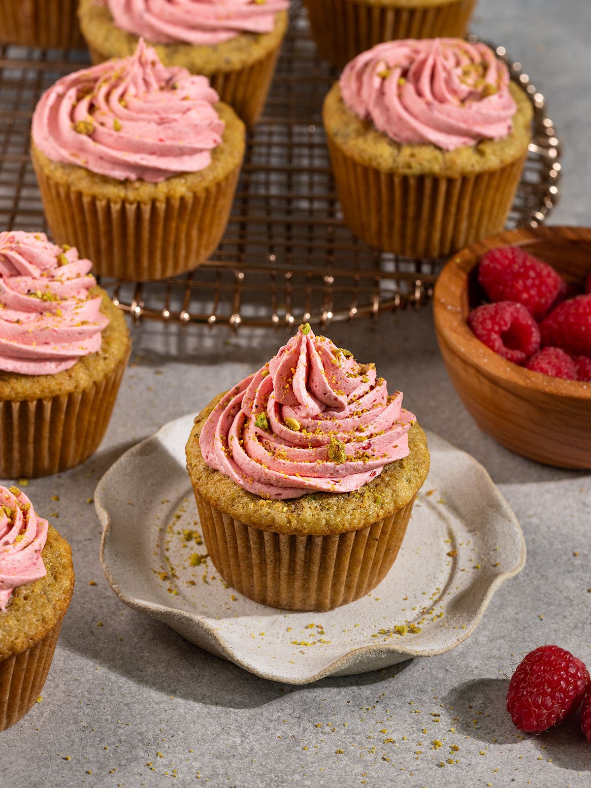 pistachio cupcakes topped with raspberry frosting and crushed pistachios on plates and a wire rack.