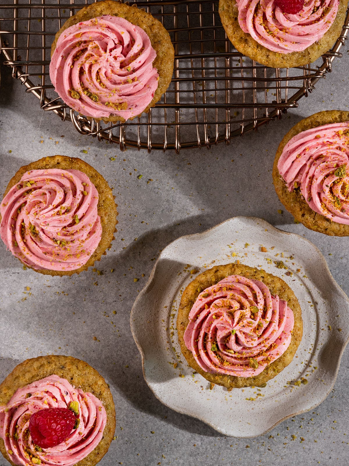 overhead view of cupcakes topped with pink raspberry frosting, crushed pistachios, and some with fresh raspberries on top.