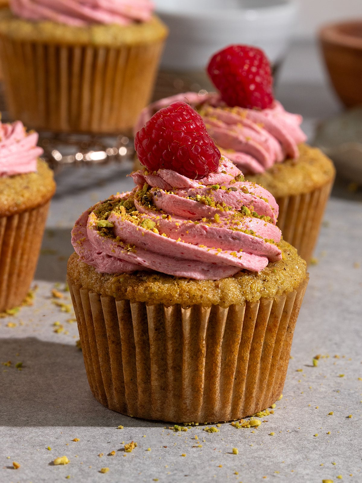 pistachio cupcake topped with raspberry frosting, crushed pistachios, and fresh raspberries with more cupcakes behind.