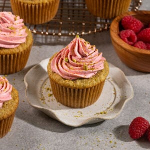 pistachio cupcakes topped with raspberry frosting and crushed pistachios on plates and a wire rack.
