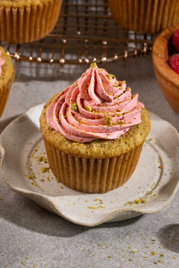 pistachio cupcakes topped with raspberry frosting and crushed pistachios on plates and a wire rack.