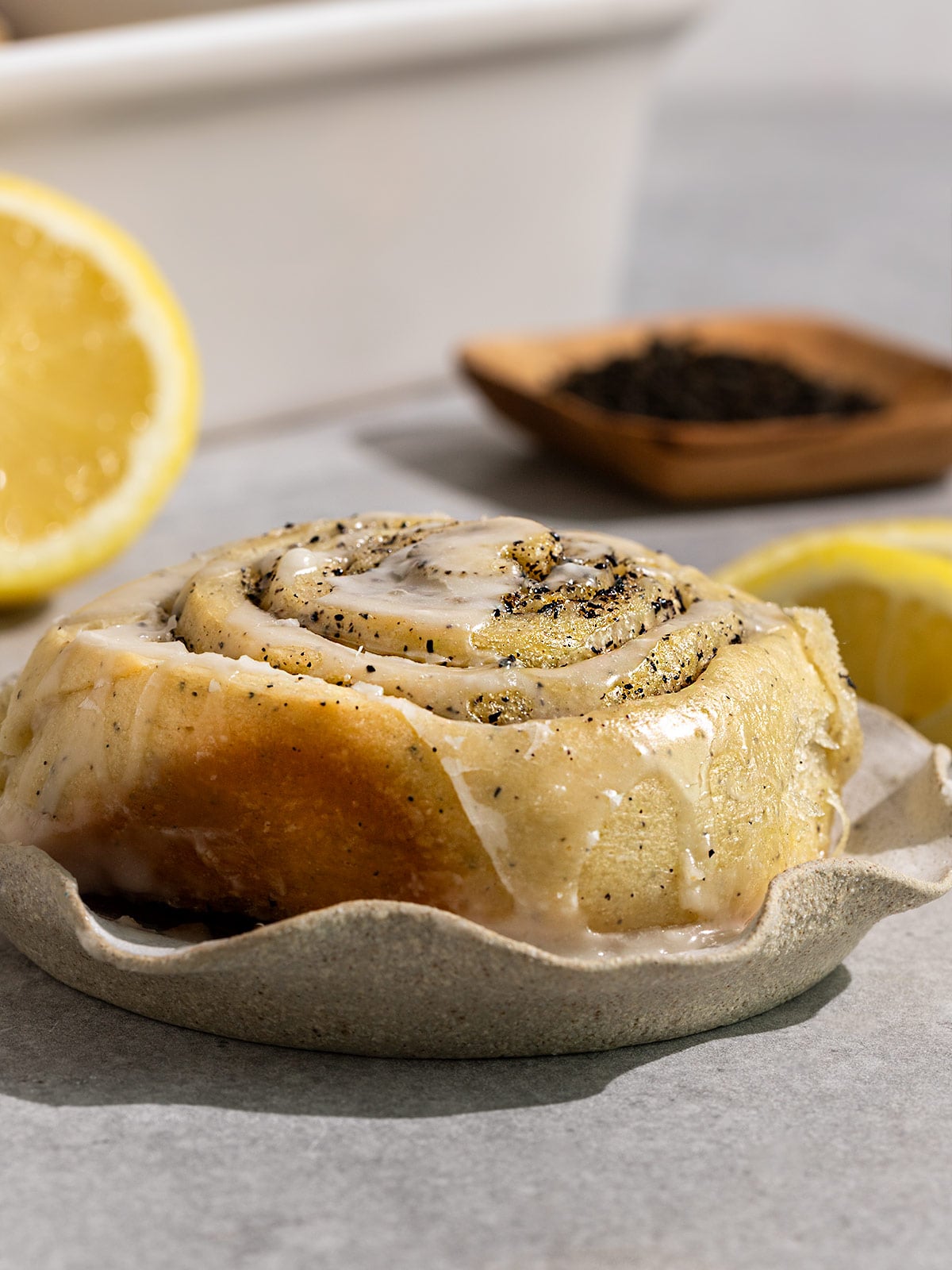 Plate toped with an earl grey lemon roll with icing with a bowl of earl grey tea leaves and lemon slices in background.