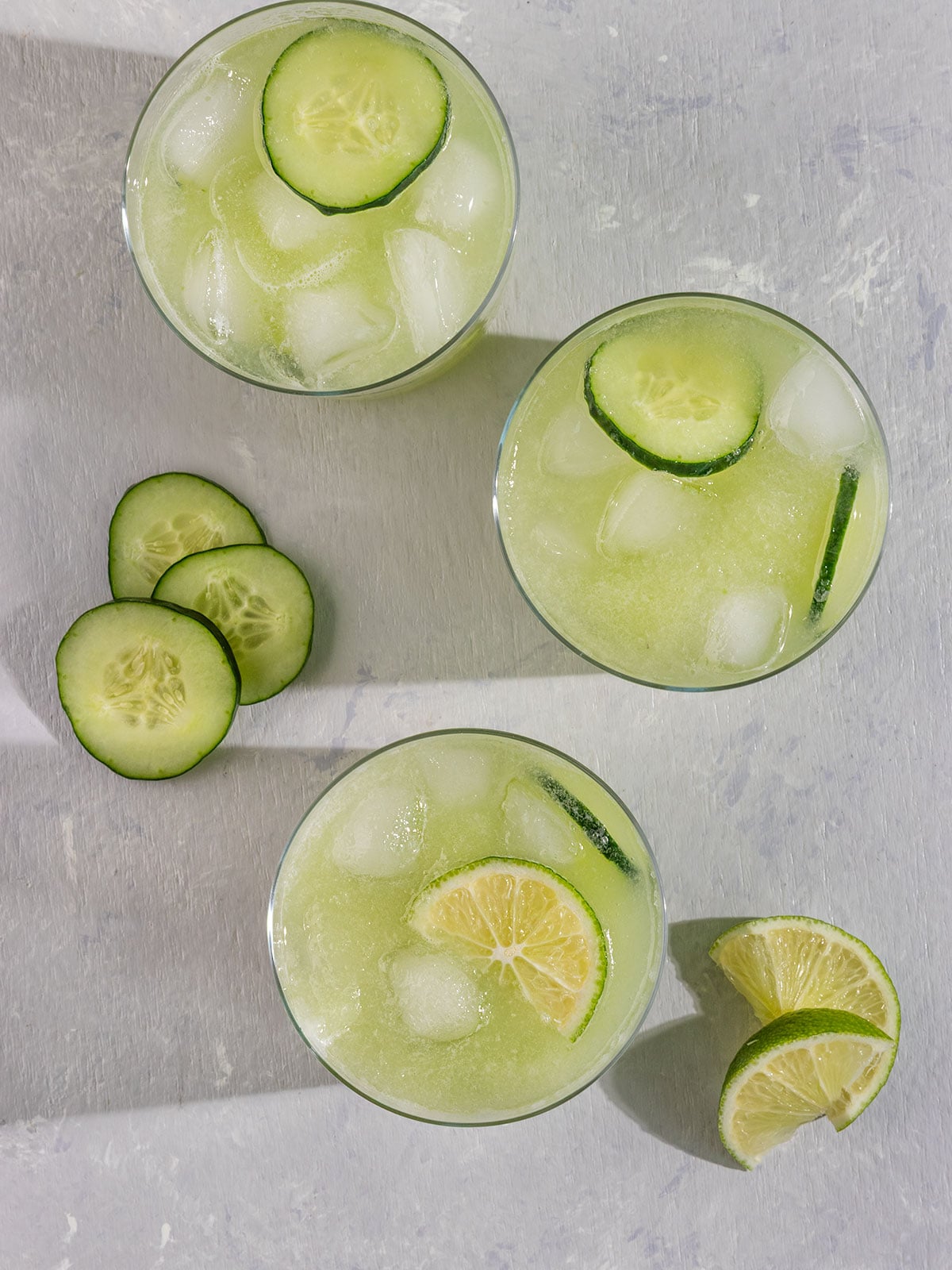 overhead view of three glasses of green juice with cucumber slices and lime wedges.