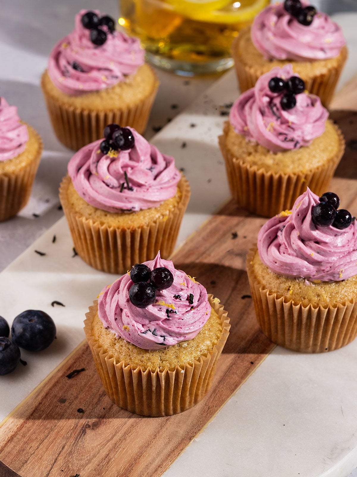 wooden and marble board topped with many earl grey cupcakes topped with purple blueberry frosting and fresh blueberries.
