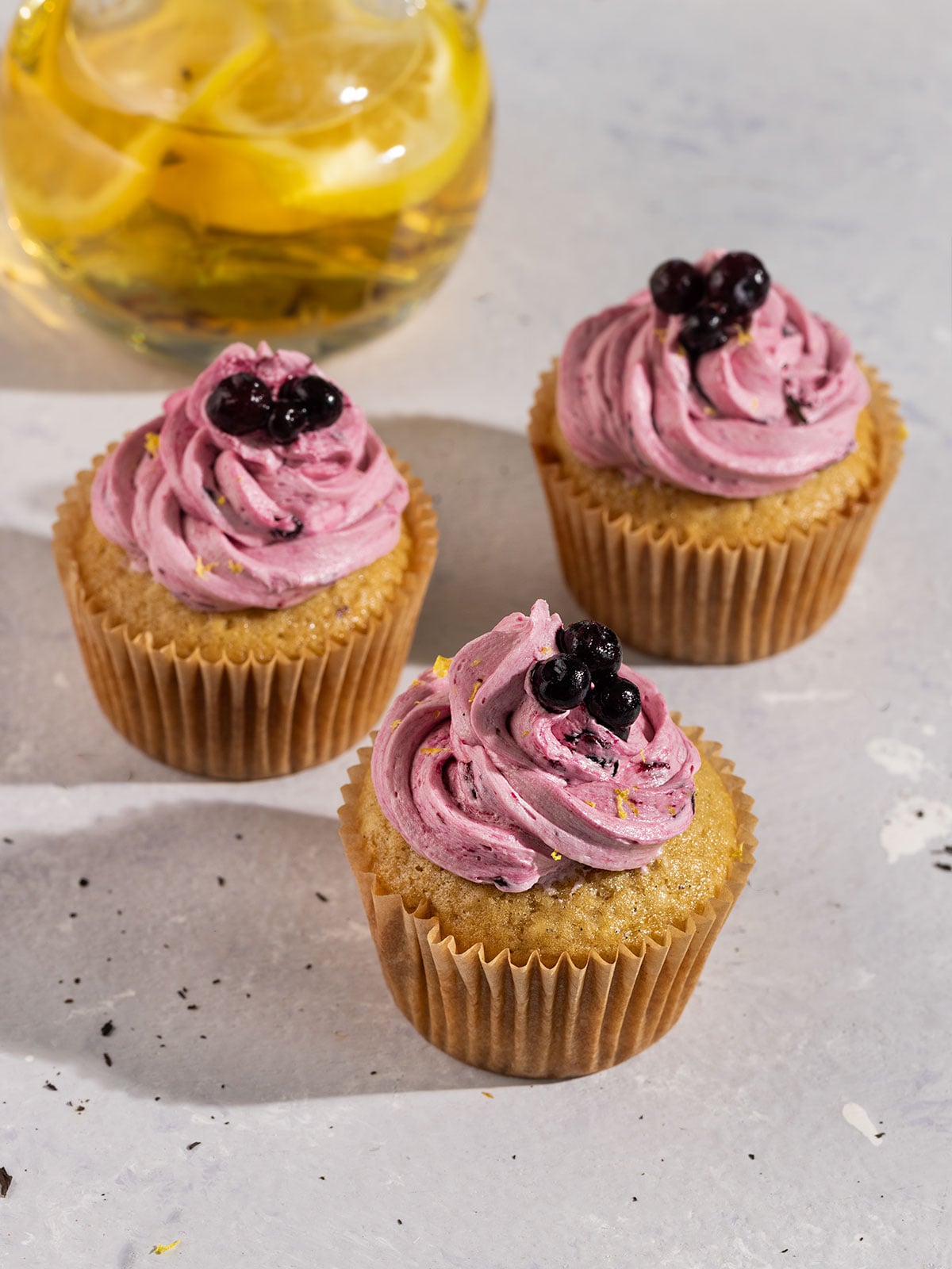 three earl grey cupcakes topped with purple blueberry frosting and fresh blueberries with a pot of earl grey lemon tea.