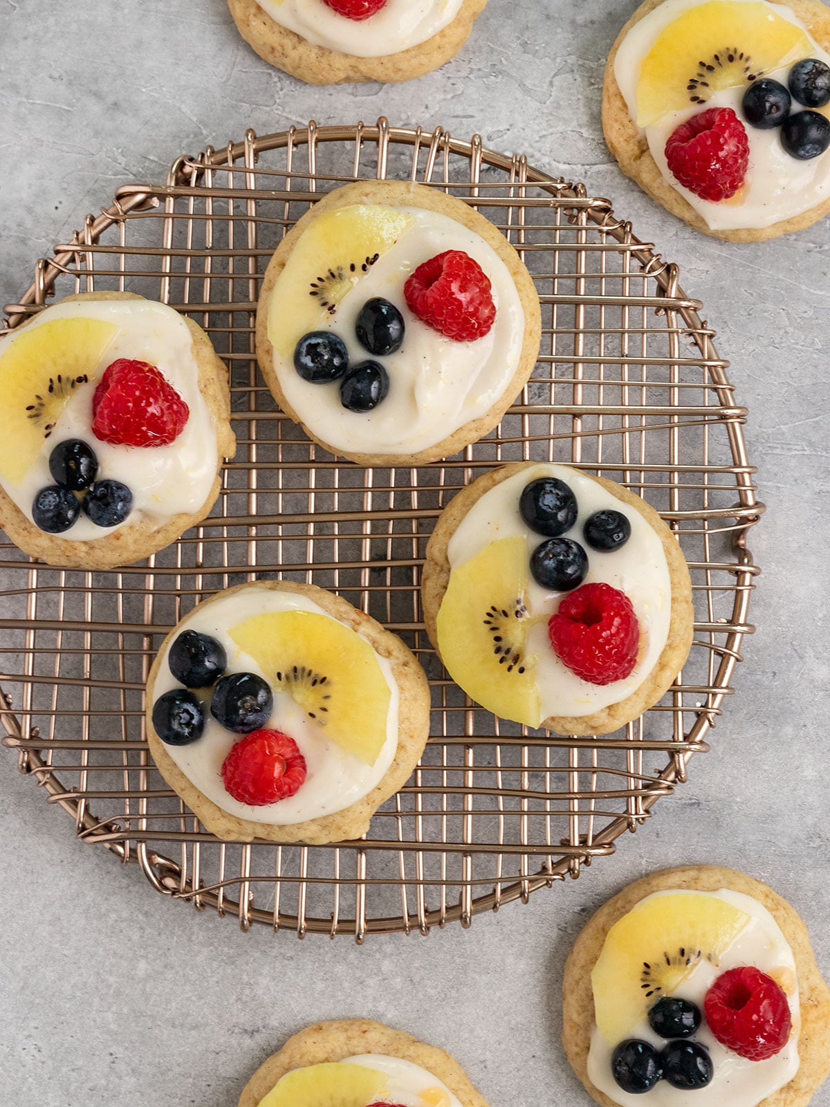 overhead shot of cookies on rose gold circle cooling rack topped with cream cheese frosting, sliced kiwi, raspberries, blueberries, and apricot jam.