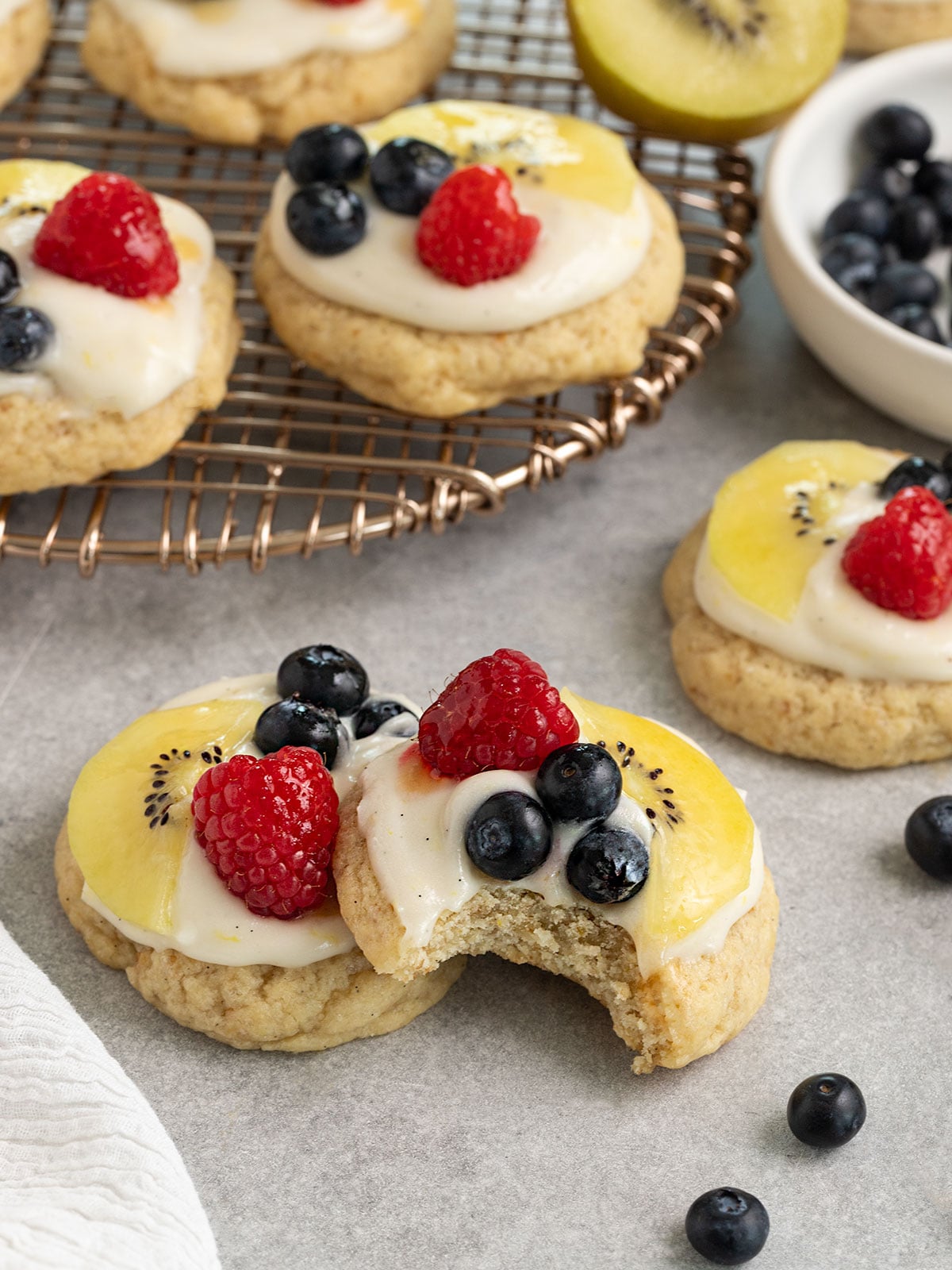 two cookies topped with cream cheese frosting, sliced kiwi, raspberries, blueberries, and apricot glaze with one cookie having a bite taken out in front of a rack with more cookies.