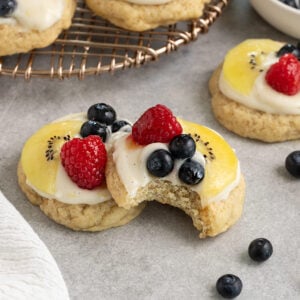 two cookies topped with cream cheese frosting, sliced kiwi, raspberries, blueberries, and apricot glaze with one cookie having a bite taken out.