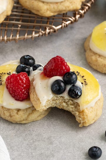 two cookies topped with cream cheese frosting, sliced kiwi, raspberries, blueberries, and apricot glaze with one cookie having a bite taken out.