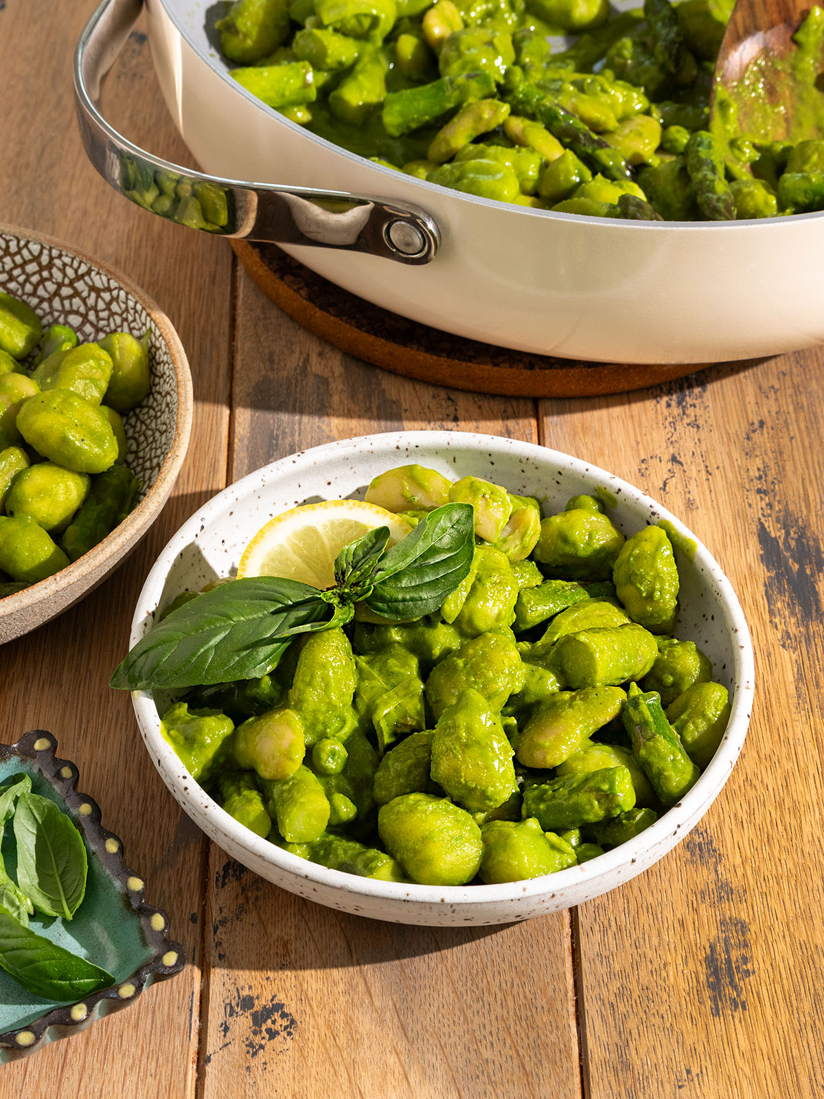 two bowls filled with gnocchi, asparagus, peas, leeks, and butter beans covered in a green pesto sauce with fresh basil on top alongside a pan of more gnocchi.