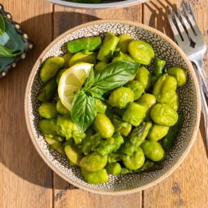 bowl filled with gnocchi, asparagus, peas, leeks, and butter beans covered in a green pesto sauce with fresh basil on top with fork on the side.