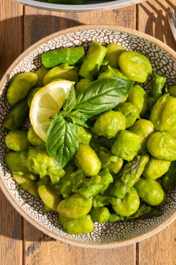 bowl filled with gnocchi, asparagus, peas, leeks, and butter beans covered in a green pesto sauce with fresh basil on top with fork on the side.
