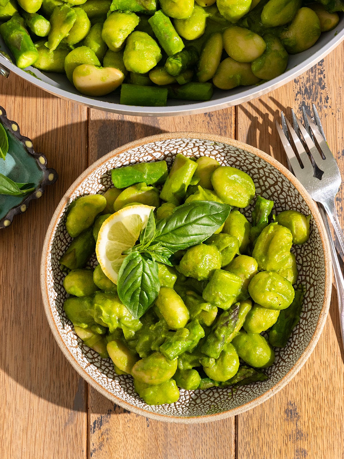 bowl filled with gnocchi, asparagus, peas, leeks, and butter beans covered in a green pesto sauce with fresh basil on top alongside a pan of more gnocchi and a fork.