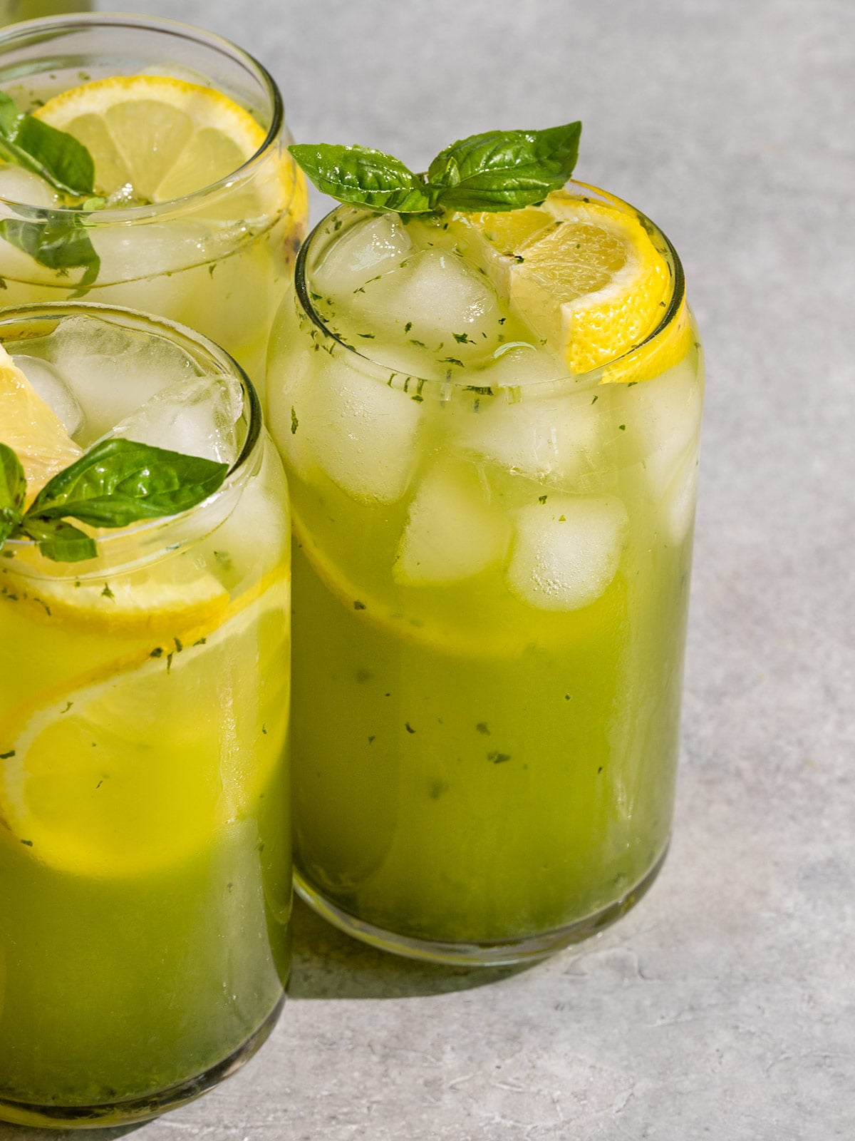 three glasses filled with a green basil lemonade, ice, slices of lemon, and a sprig of basil on top.