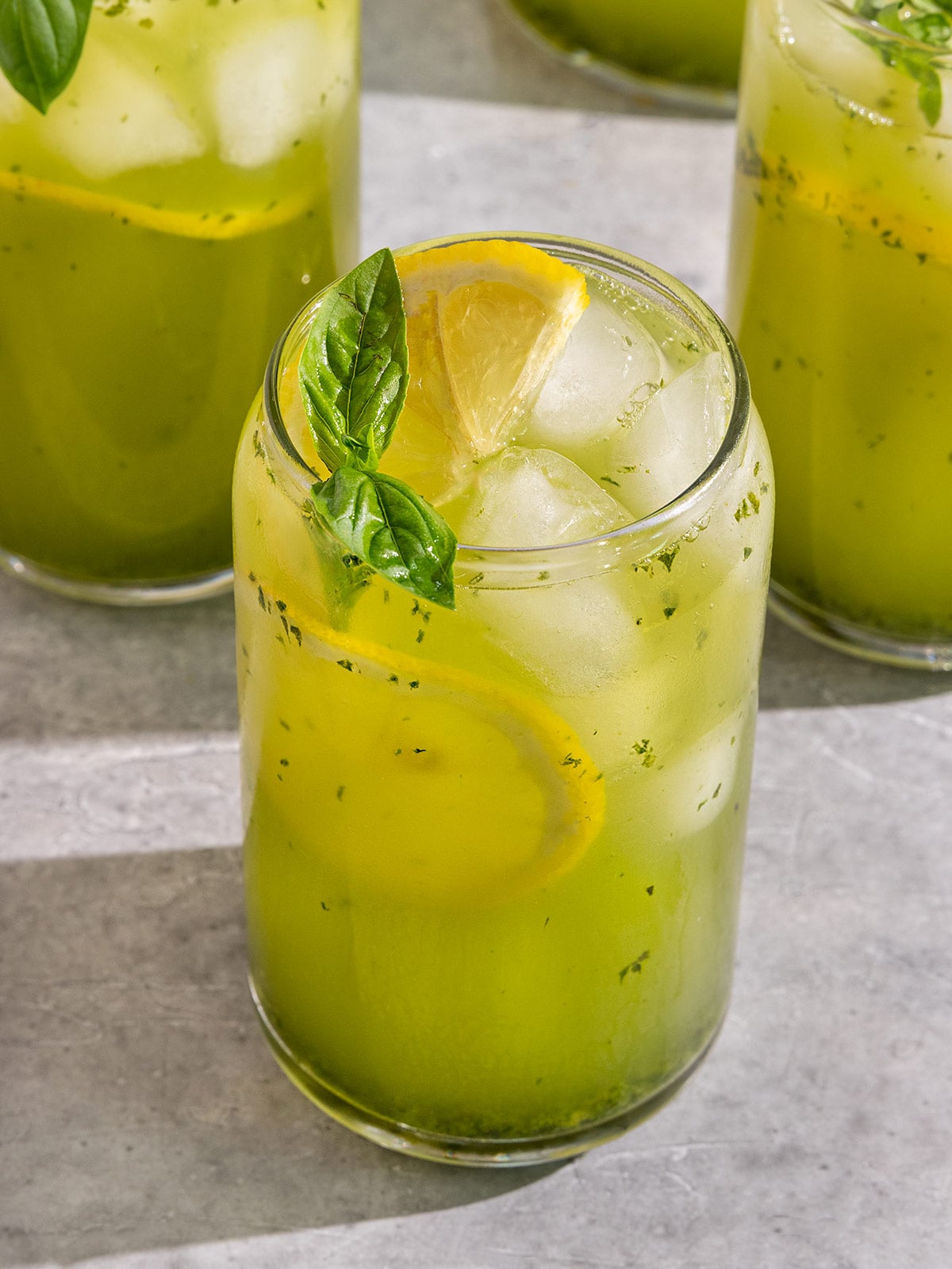 three glasses filled with a green basil lemonade, ice, slices of lemon, and a sprig of basil on top.