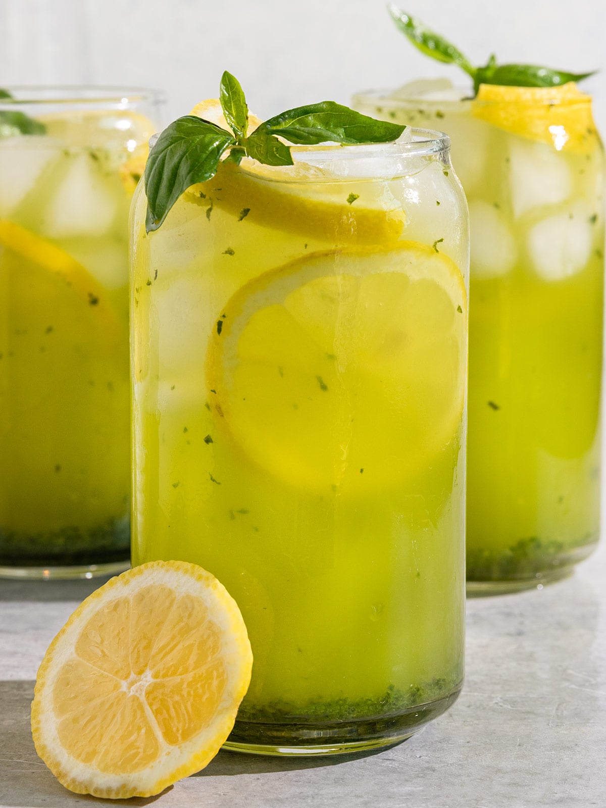 three glasses filled with a green basil lemonade, ice, slices of lemon, and a sprig of basil on top.