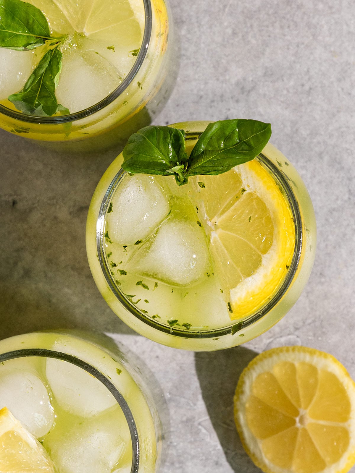 overhead view of three glasses with green basil lemonade, wedges of fresh lemon, and sprigs on basil inside.