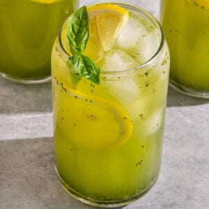 glass filled with a green basil lemonade, slices of lemon, and a sprig of basil on top.