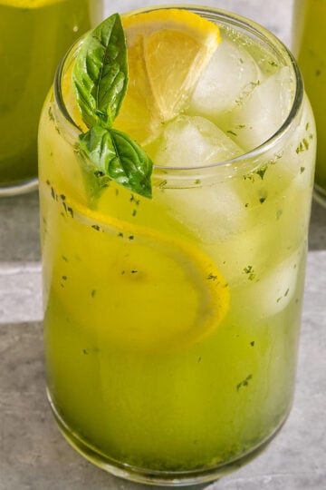 glass filled with a green basil lemonade, slices of lemon, and a sprig of basil on top.