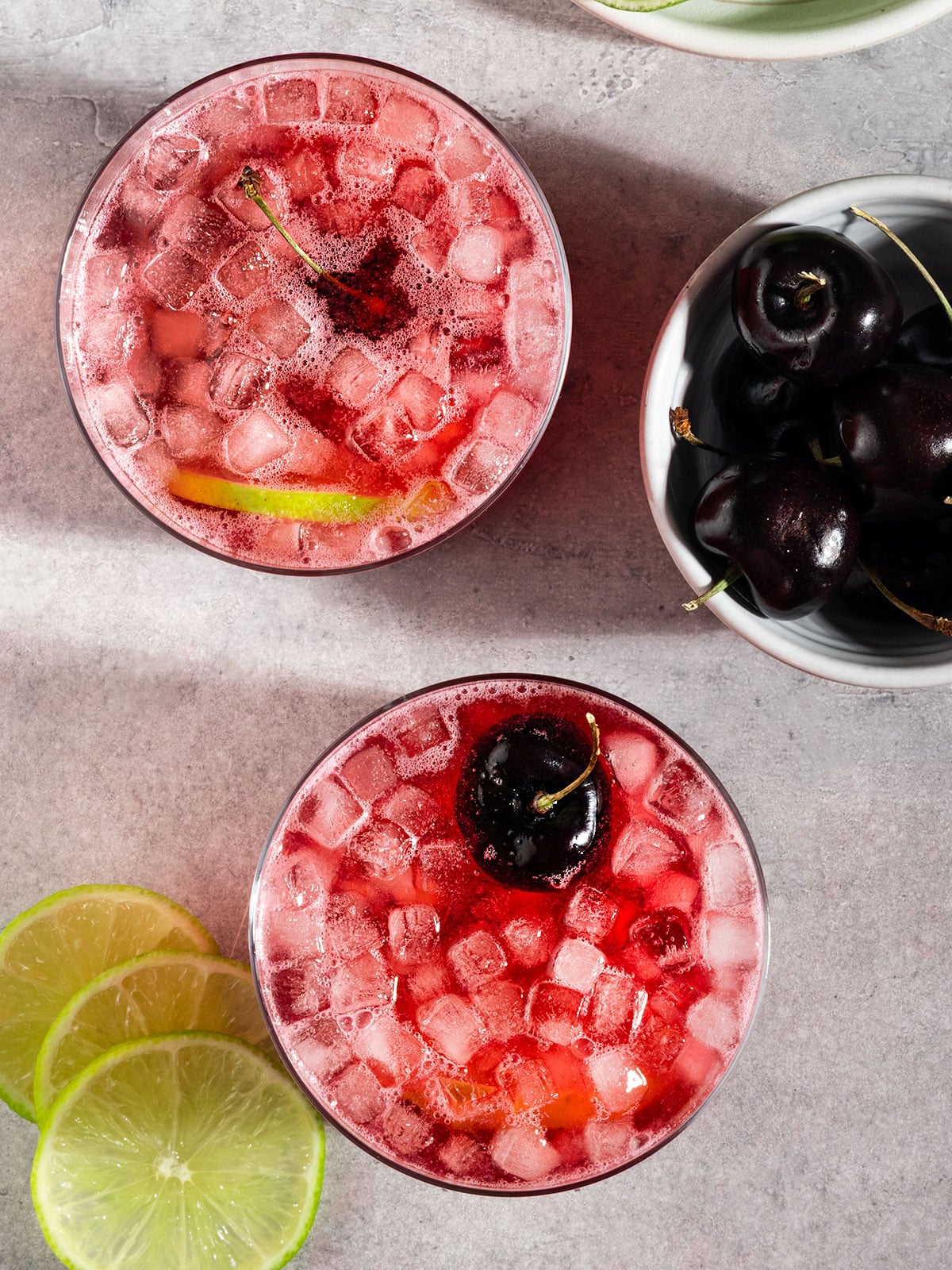 two glasses of cherry lime soda with ice, fresh lime slice, and cherries.