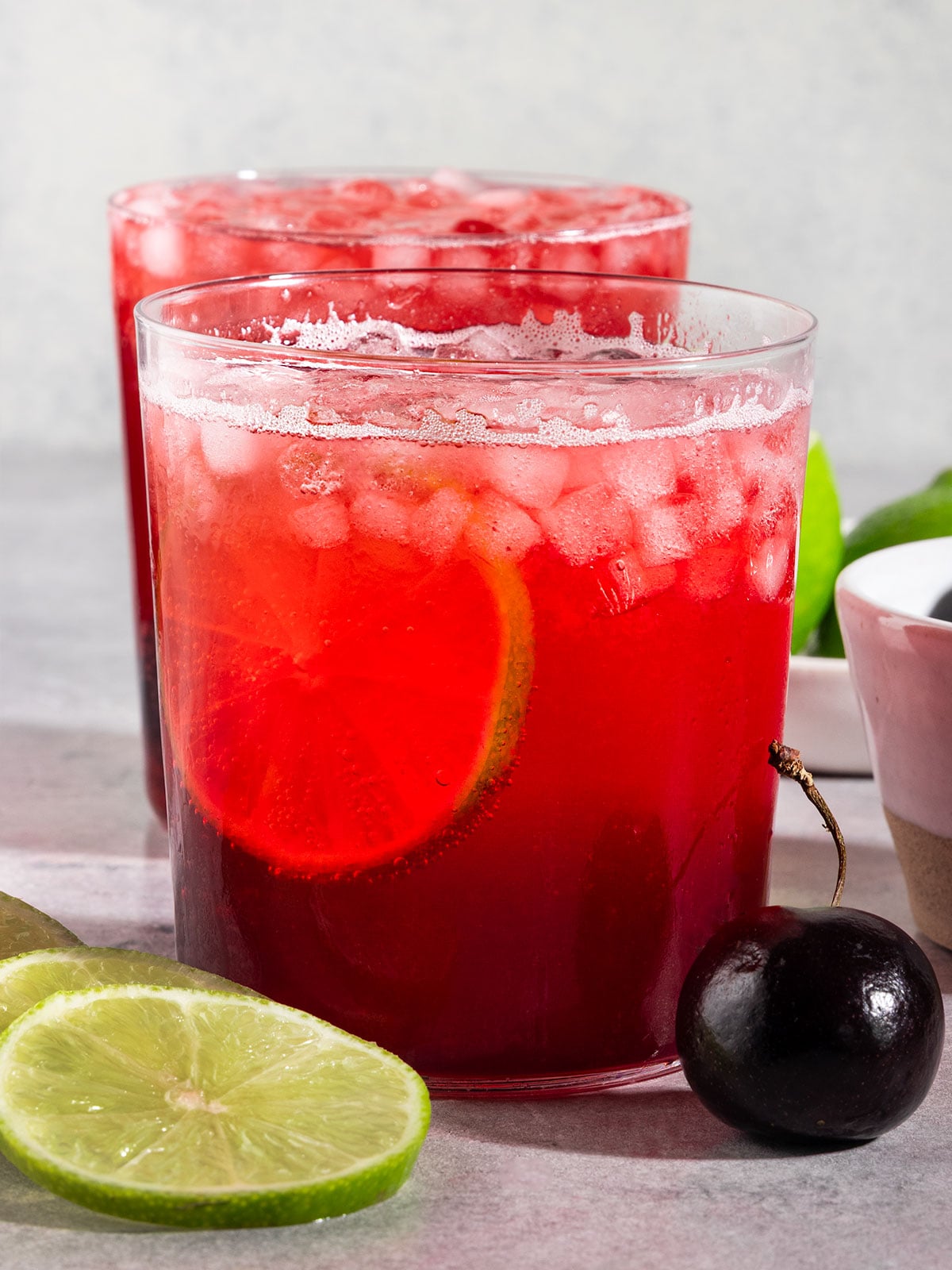 two glasses of cherry lime soda with ice, fresh lime slice, and cherries.