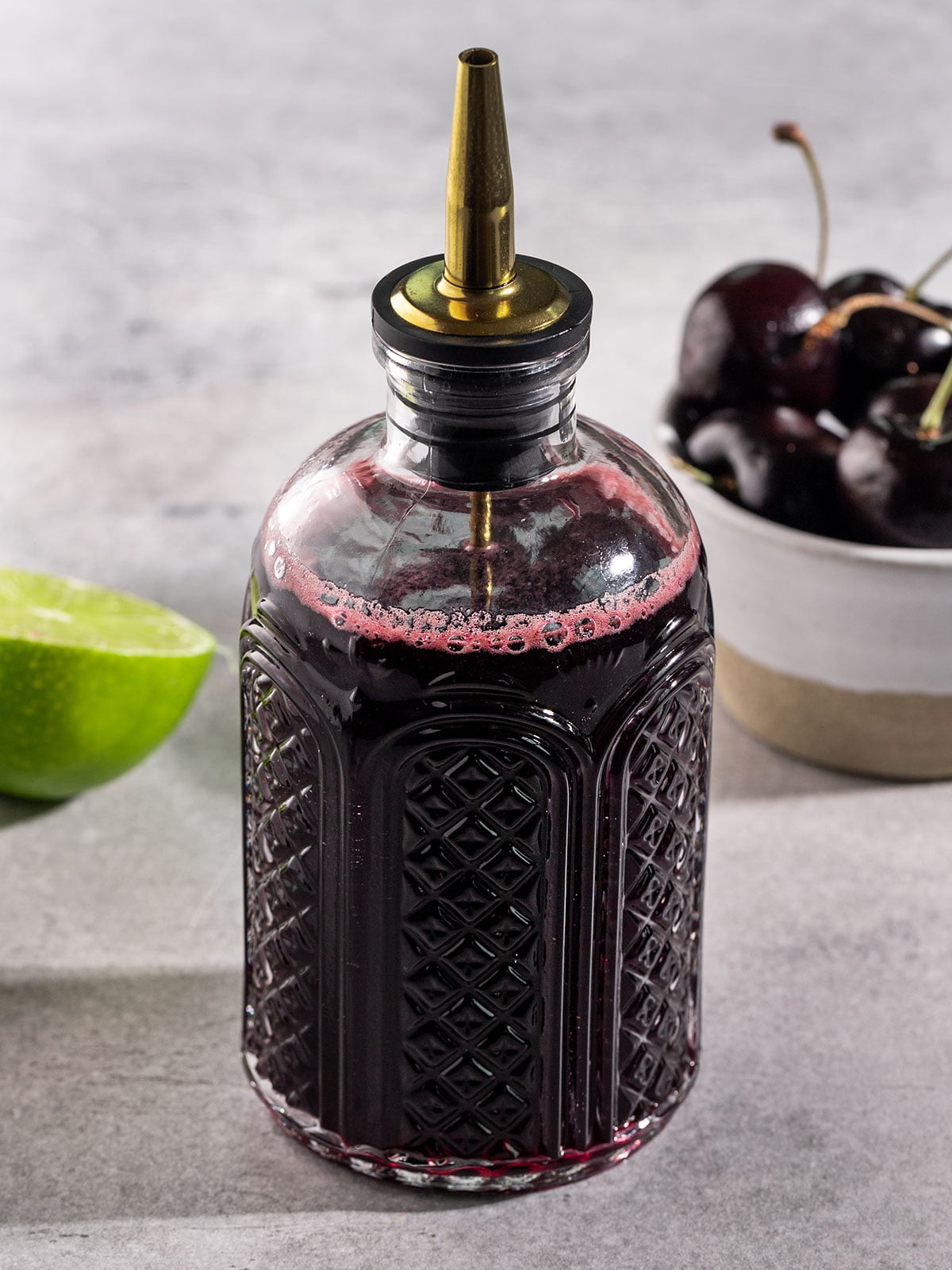 glass bottle filled with cherry syrup alongside bowl of cherries and half a lime.