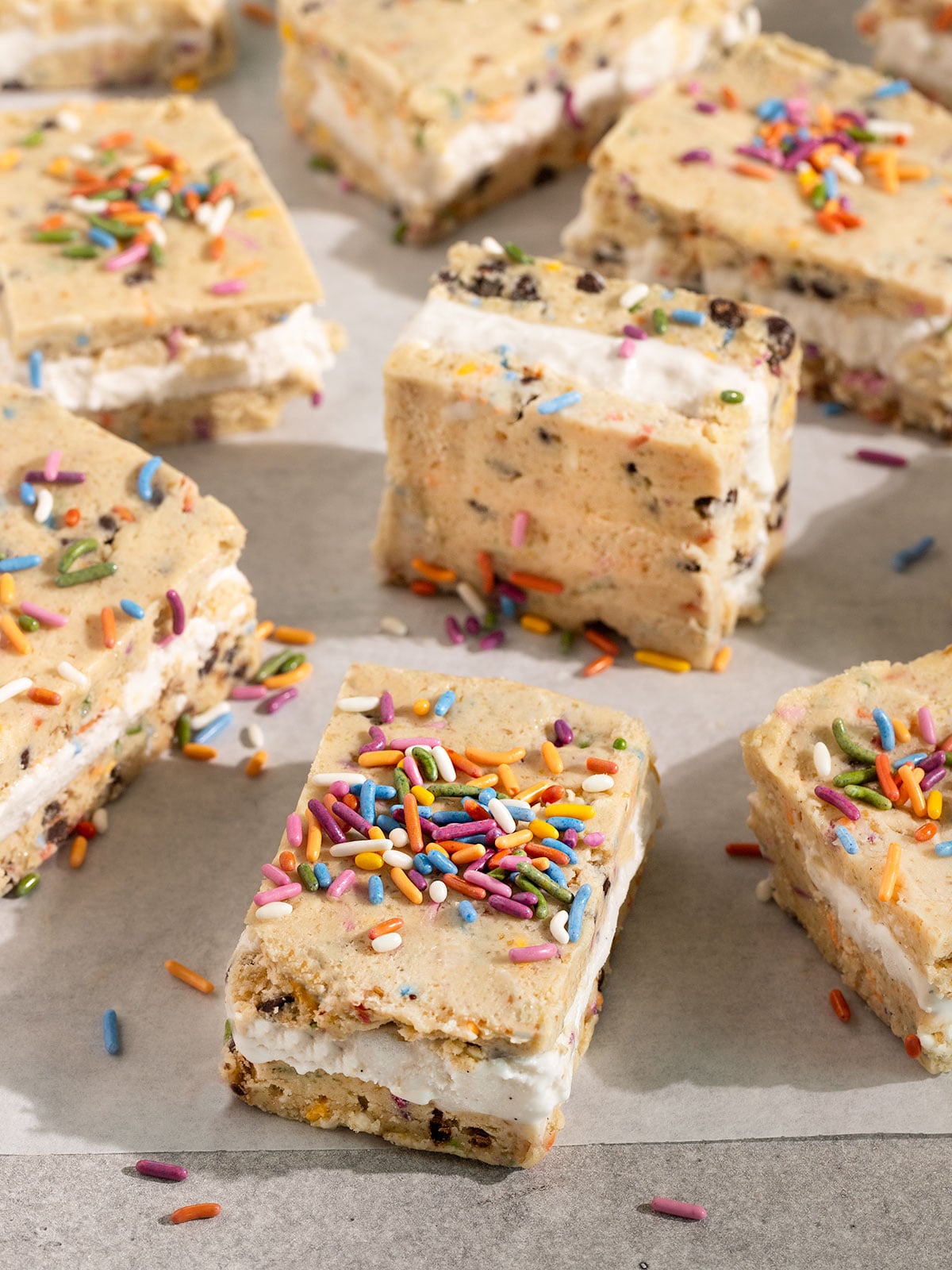 white parchment paper topped with squares of cookie dough ice cream sandwiches filled with vanilla ice cream and topped with rainbow sprinkles.
