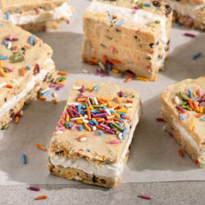 white parchment paper topped with squares of cookie dough ice cream sandwiches filled with vanilla ice cream and topped with rainbow sprinkles.