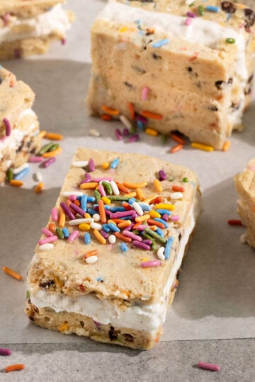 white parchment paper topped with squares of cookie dough ice cream sandwiches filled with vanilla ice cream and topped with rainbow sprinkles.