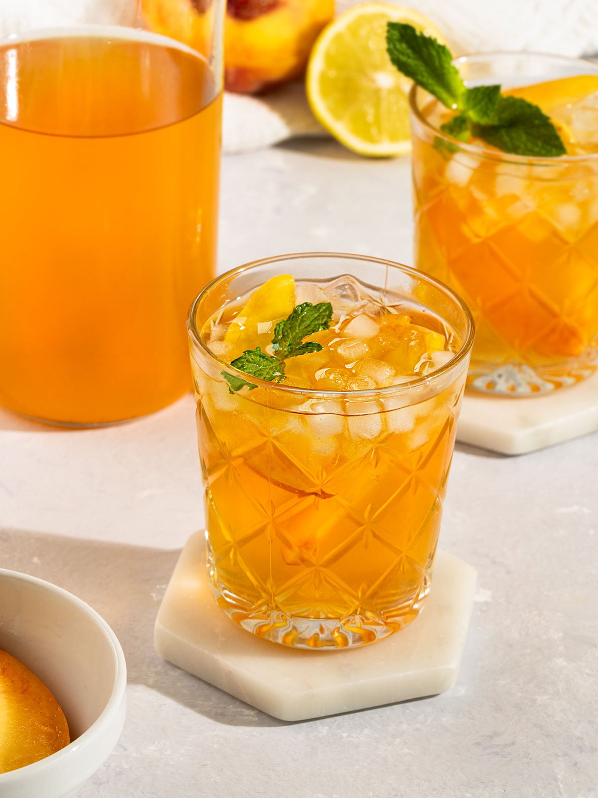 two glasses of peach tea filled with ice, fresh peach slices, and topped with sprig of mint alongside a tall glass bottle of tea.