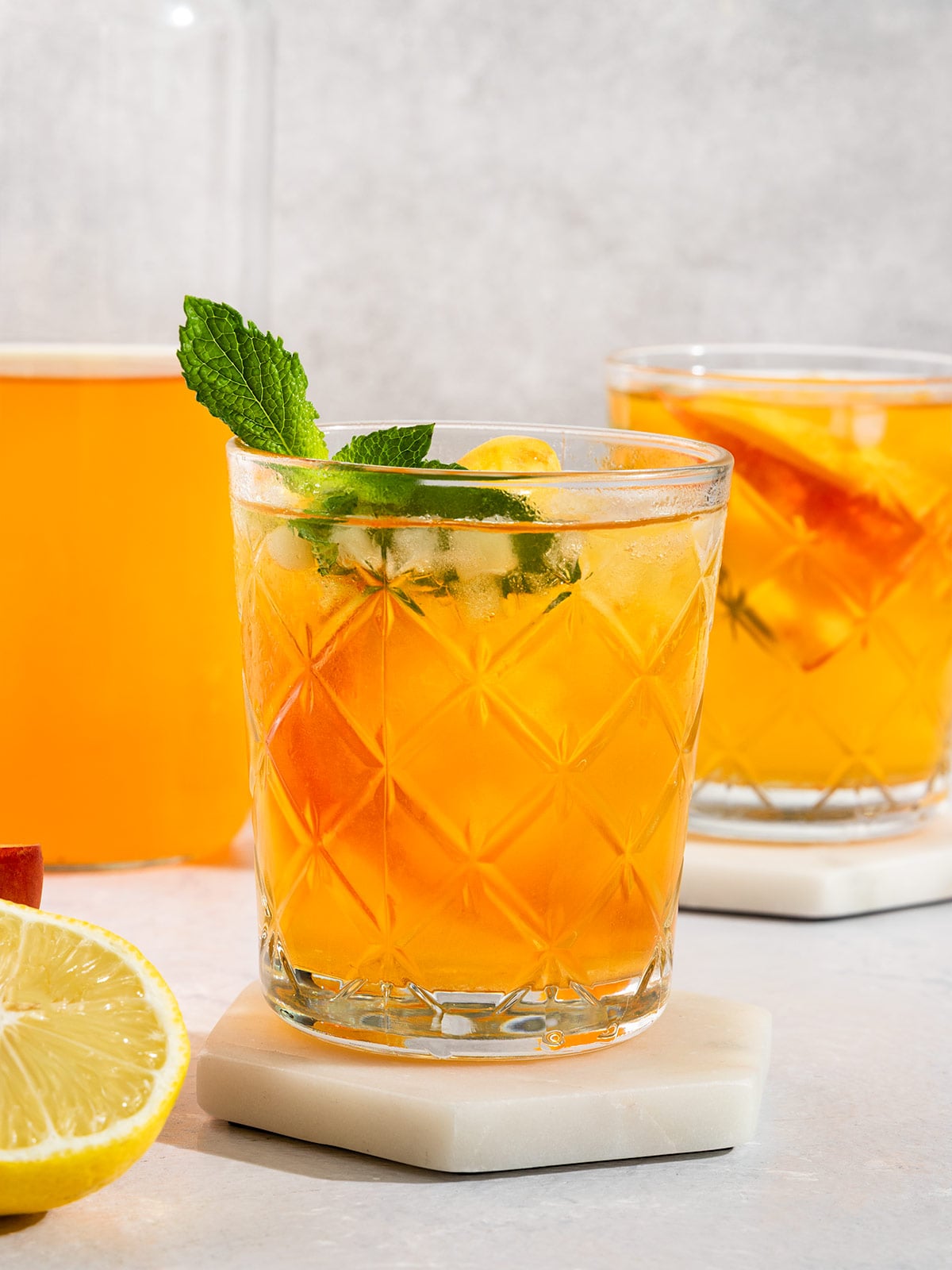 two glasses of peach tea filled with ice, fresh peach slices, and topped with sprig of mint alongside a tall glass bottle of tea.