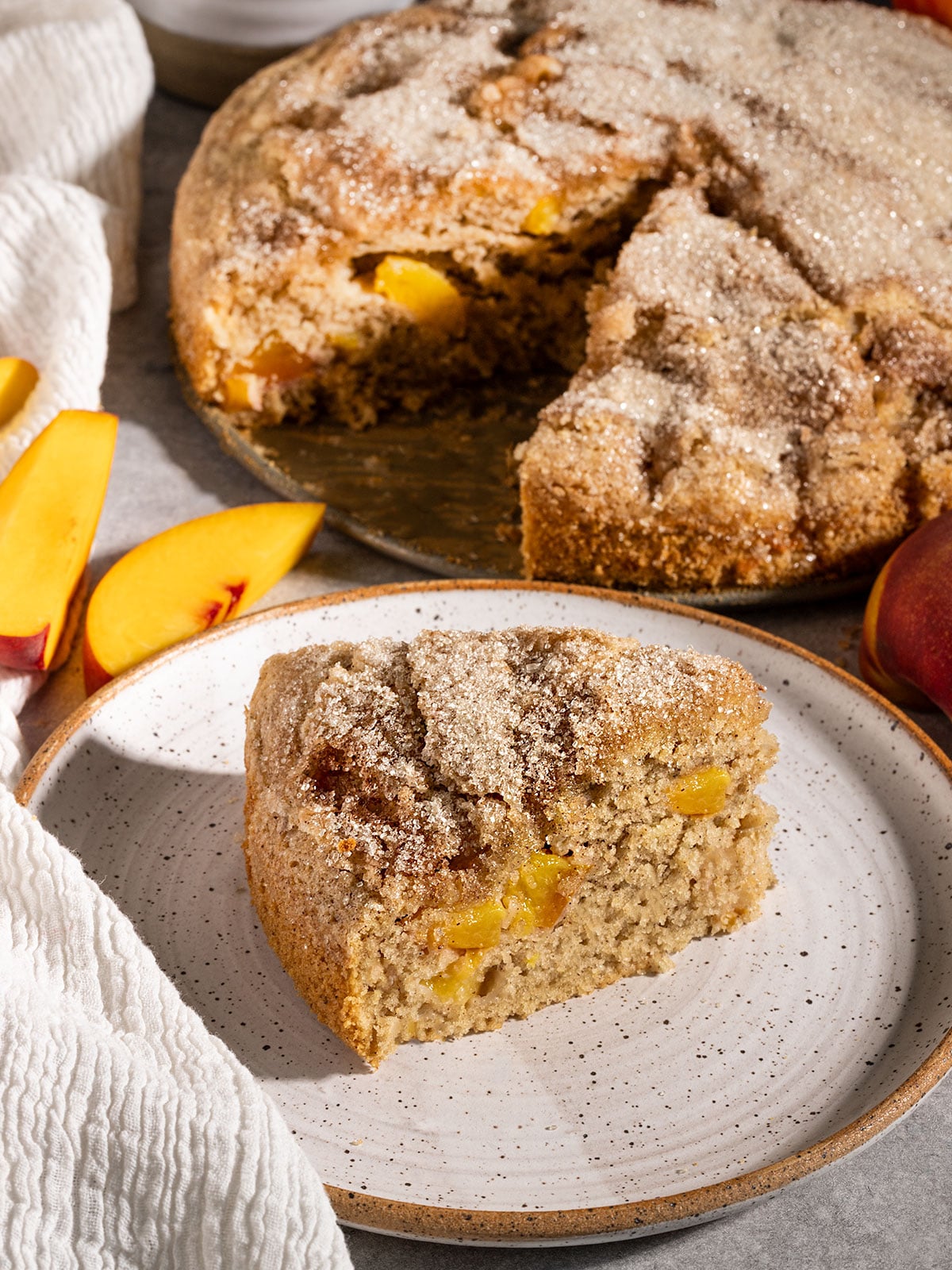 slice of cake on a plate with fresh peaches inside and a cinnamon sugar topping in front of the remaining cake.