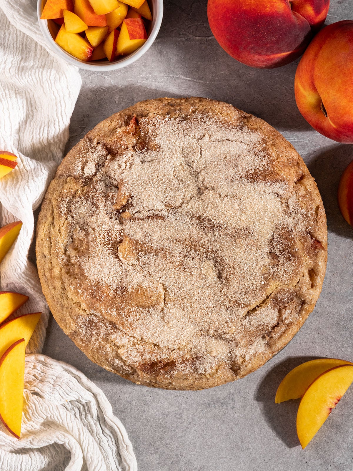 circular cake with cinnamon sugar topping surrounded by fresh peaches.