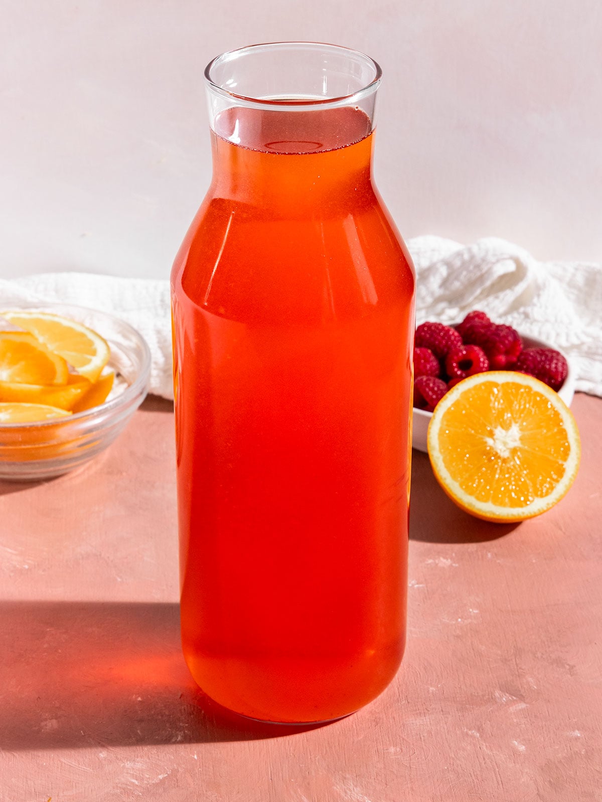 tall glass bottle of pink/red tea with bowl of raspberries and orange slices on side.