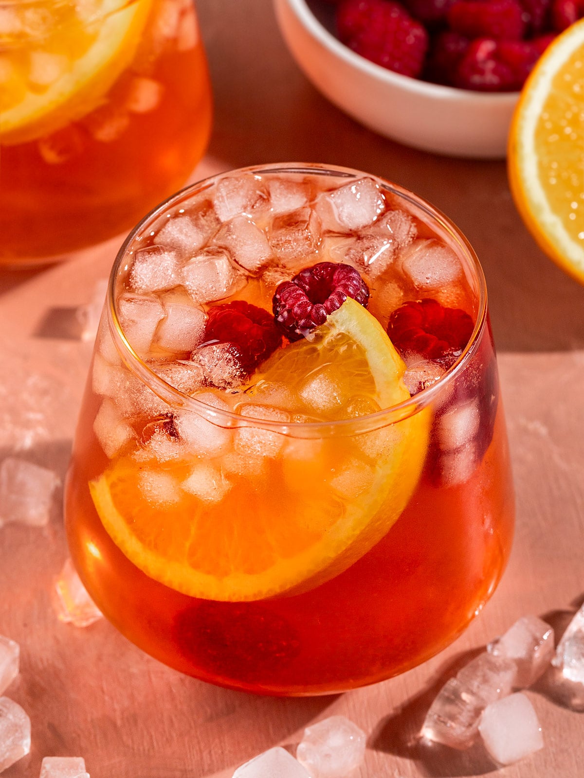 glass of pink iced tea with fresh orange slice, raspberries, and small square ice inside.