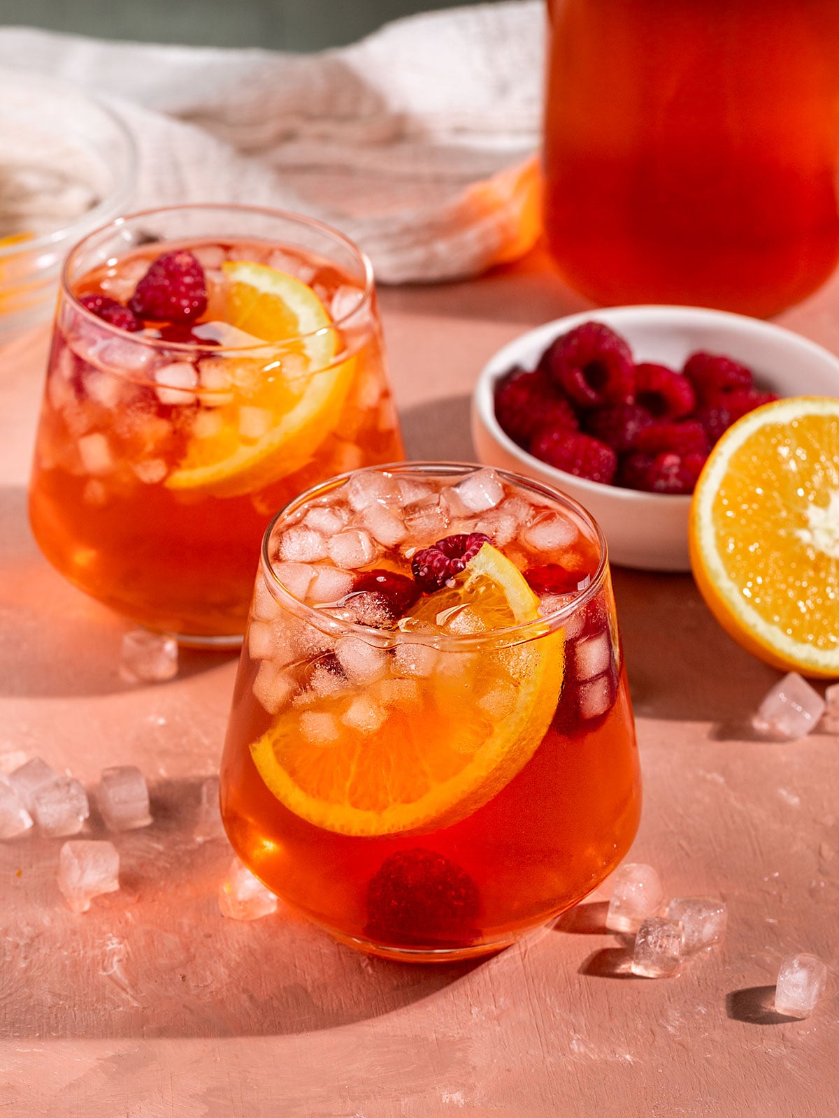 two glasses of pink/red iced tea with fresh orange slice, raspberries, and small square ice inside with bowl of raspberries and half an orange on the side.