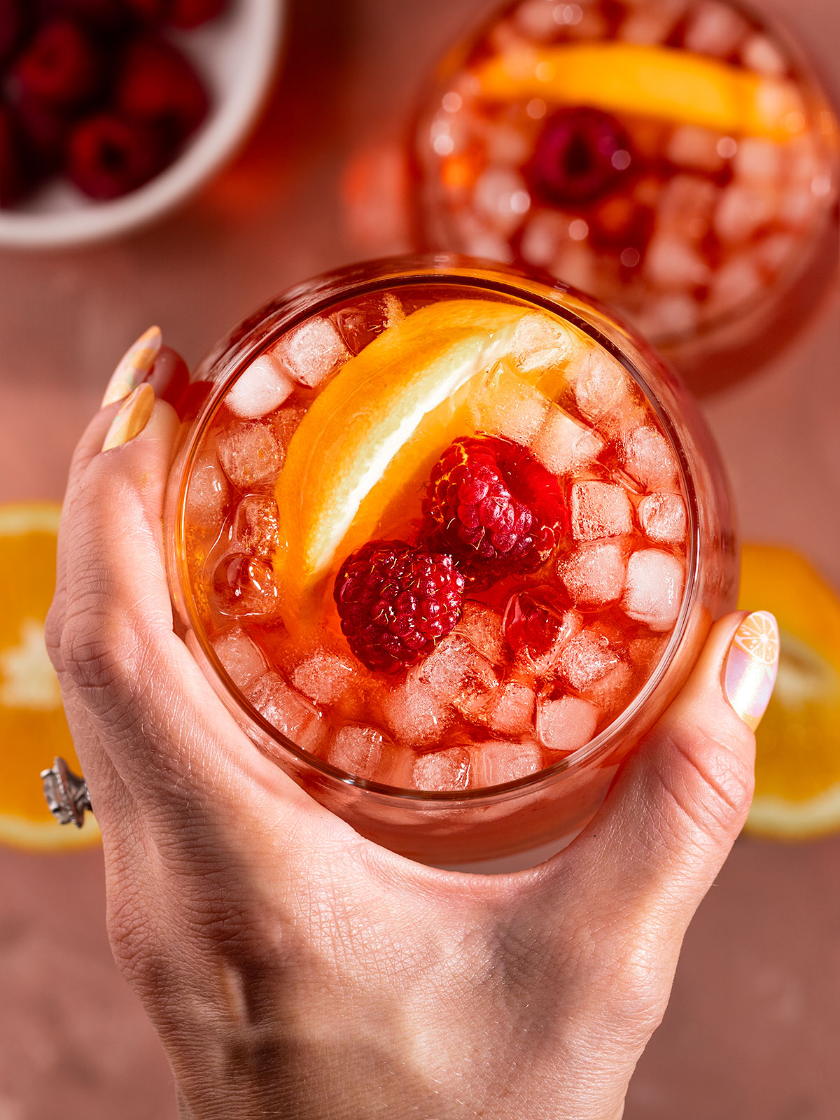 hand holding a glass filled with tea, ice, raspberries, and orange slices.