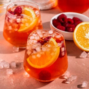 two glasses of pink/red iced tea with fresh orange slice, raspberries, and small square ice inside with bowl of raspberries and half an orange on the side.