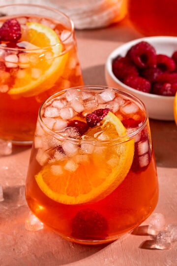 two glasses of pink/red iced tea with fresh orange slice, raspberries, and small square ice inside with bowl of raspberries and half an orange on the side.