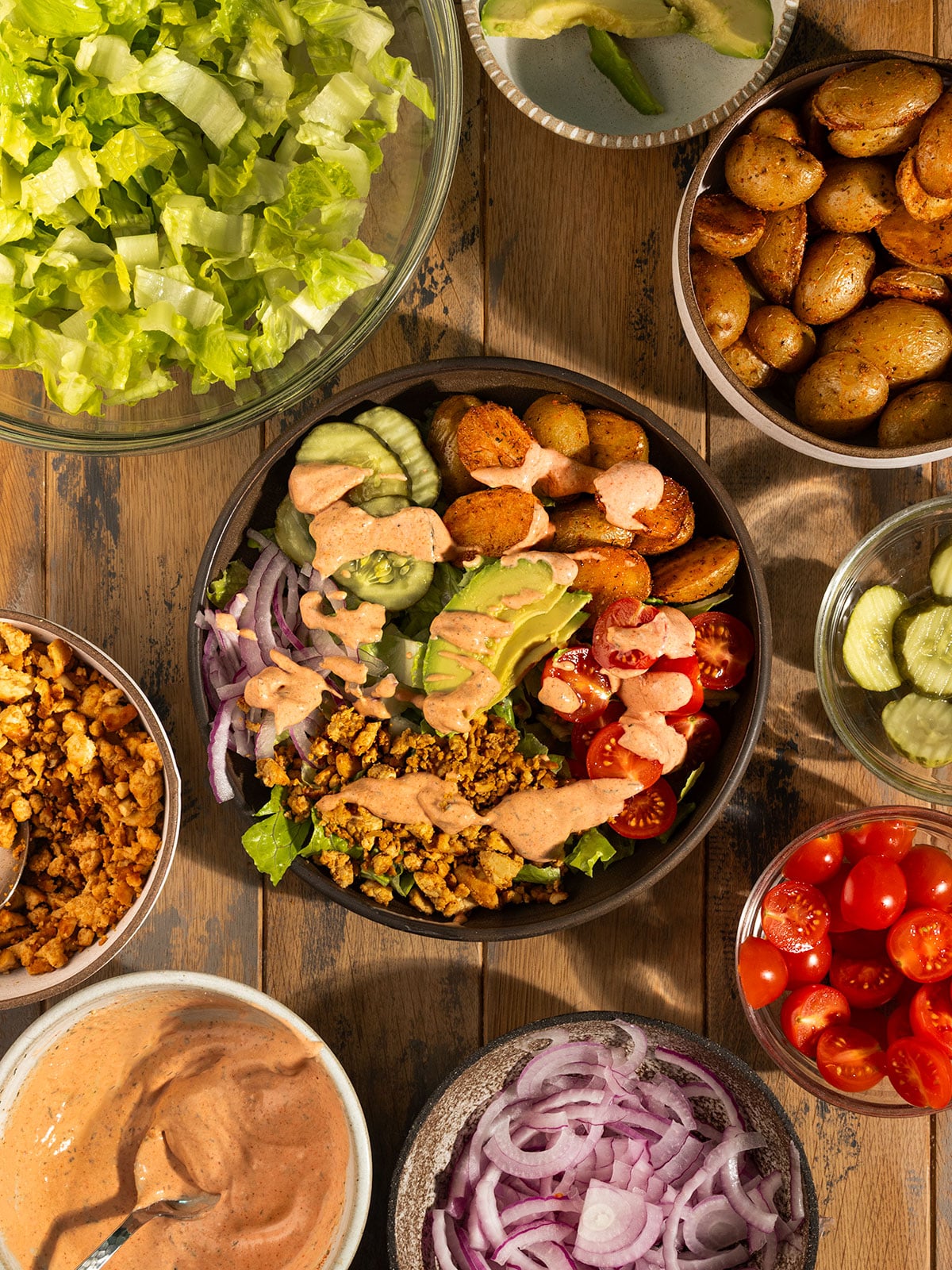 burger bowl filled with romaine lettuce, roasted potatoes, tomatoes, sliced red onion, pickles, avocado, tofu crumbles, and topped with a burger sauce with bowls of extra toppings on sides.