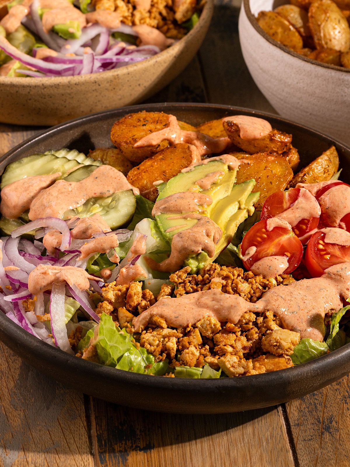 bowl filled with romaine lettuce, sliced red onions, sliced cherry tomatoes, avocado, tofu crumbles, roasted potatoes, pickles, and an orange burger sauce.