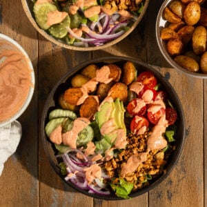 two bowls filled with romaine lettuce, roasted potatoes, tomatoes, avocado, sliced red onion, tofu crumbles, pickles, and topped with an orange burger sauce.