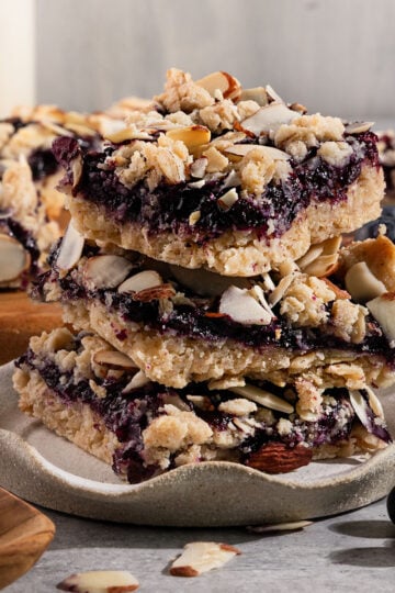 three bars with layers of cookie shortbread, blueberry filling, and almond topping stacked on a plate.